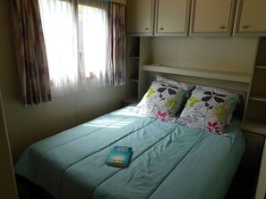 A bedroom with a bed, pillows, a book, and a window with curtains. - Cozy mobile home - 4 guests (Vesseaux)