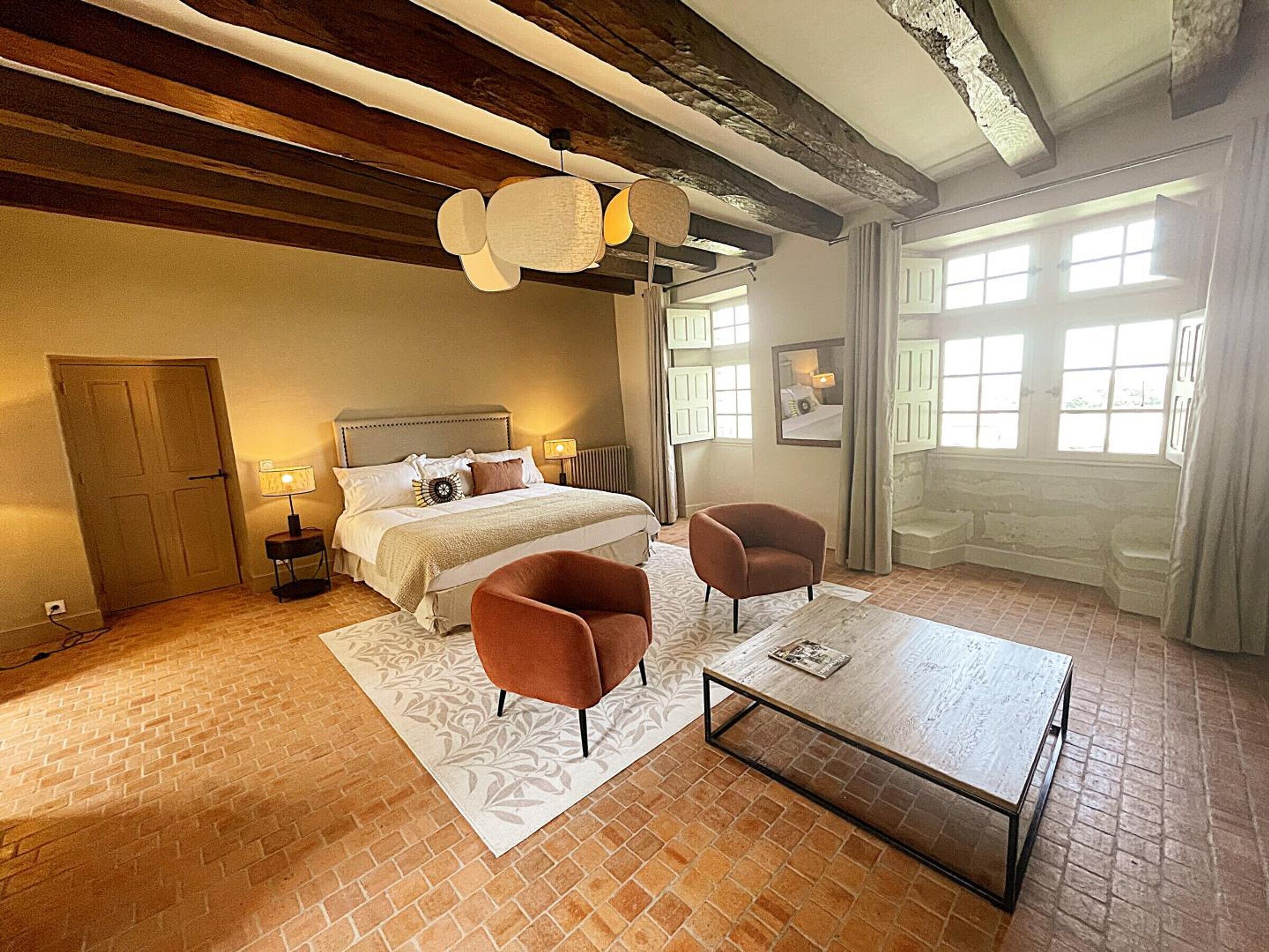 Un dormitorio con una cama, dos sillones, una mesita de centro, una chimenea y un ventanal grande.