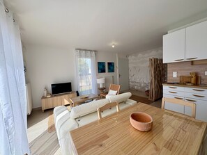 Ein modernes Wohnzimmer mit Sofa, Fernseher, Esstisch mit Schüssel und einer Küchenzeile mit Schränken.