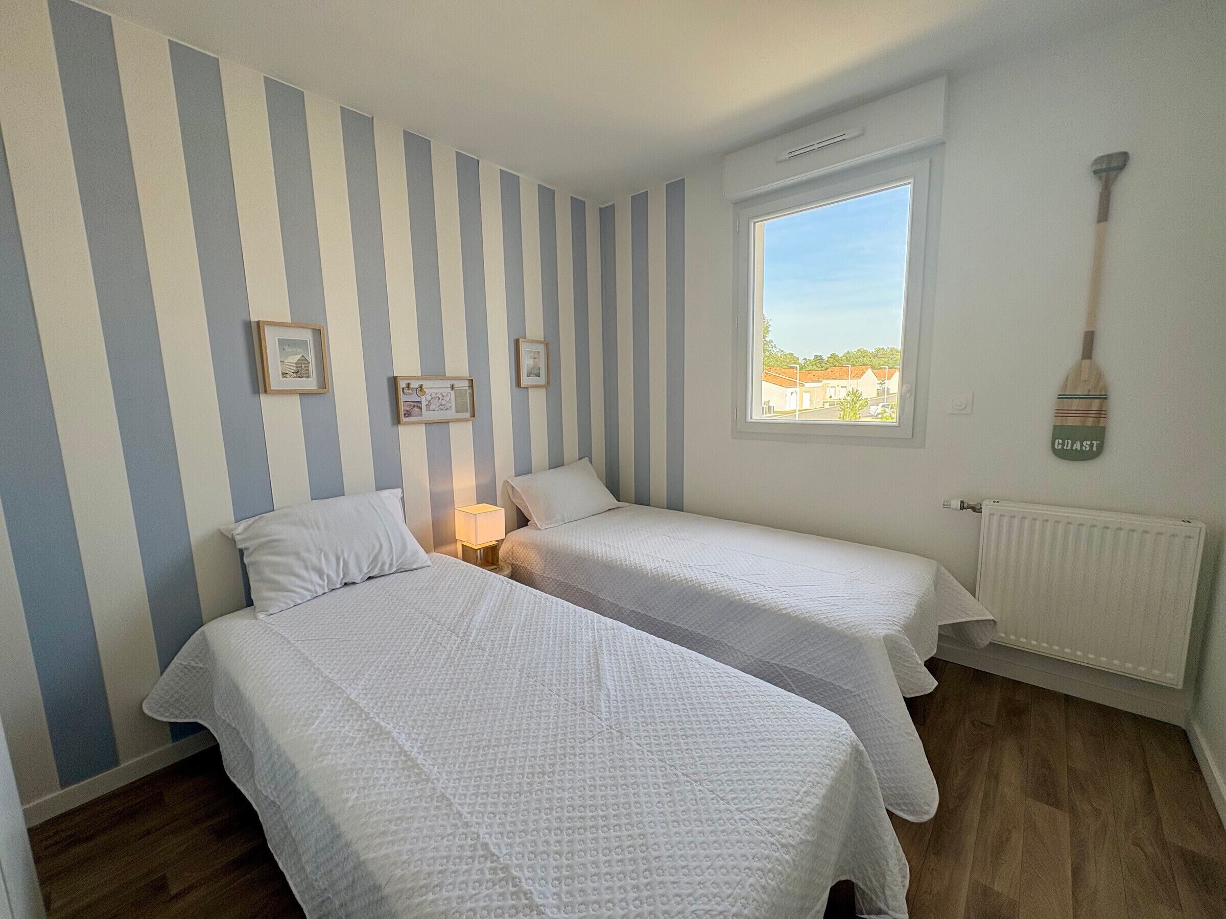 Une chambre avec deux lits, une fenêtre donnant sur un beau paysage, un radiateur et une planche de paddle accrochée au mur.