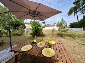 Ein Gartenhüstchen mit einem Holztisch, gelben Tellern, grünem Glas und einem braunen Sonnenschirm.