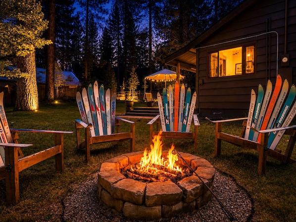Un foyer est entouré de chaises Adirondack, avec des paires de skis appuyés contre celles-ci. L’ensemble est aménagé dans un jardin, avec une cabane en bois en arrière-plan.