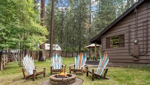 Une cabane avec un foyer entouré de fauteuils Adirondack, une terrasse en bois et une vue sur une zone boisée.