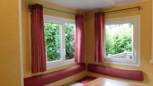Ein Zimmer mit zwei Fenstern, roten Vorhängen, einer hölzernen Bank und einer weißen Tür.