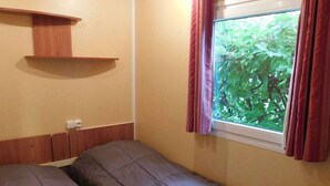 Ein Zimmer mit zwei Betten, einem Holzbodenregal, einem Fenster mit Vorhängen und einer Steckdose.