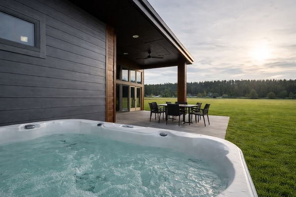 A modern house with a hot tub, a patio with a table and chairs, and a view of a grassy field and trees.