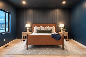 A modern bedroom with a large bed, two bedside tables with lamps, a view through a window, and a dark wall with vertical lines.