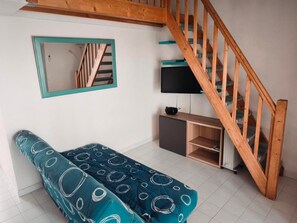 A room with a wooden staircase, a sofa with a teal and white pattern, a TV on a wooden stand, and a mirror on the wall. - T3 with AC, sea view, parking - 6 guests (Gruissan)