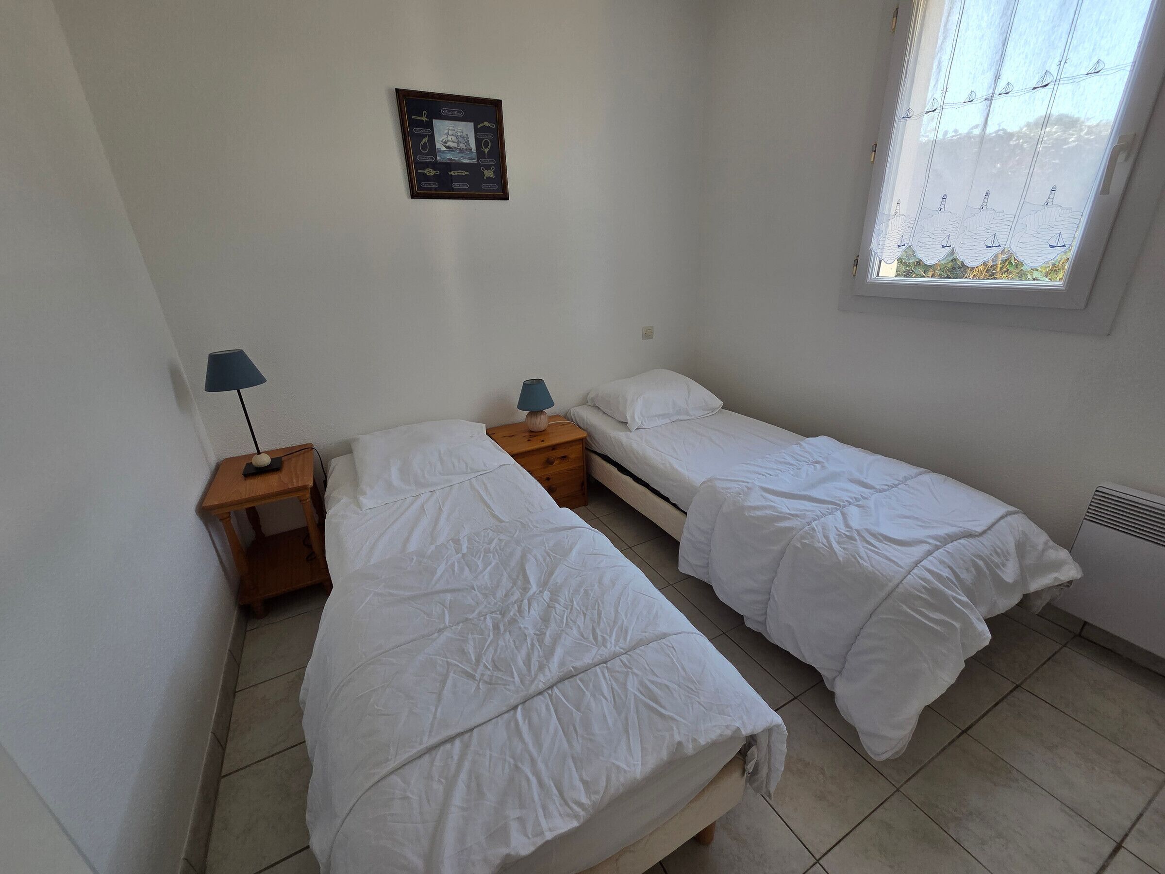 A small, clean room with two single beds, a nightstand with a lamp, a framed picture on the wall, and a window with a view.