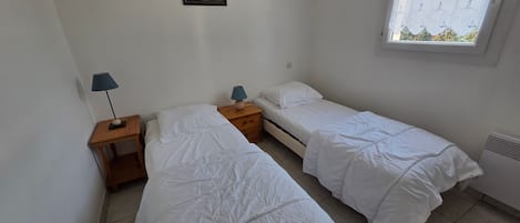 A small, clean room with two single beds, a nightstand with a lamp, a framed picture on the wall, and a window with a view.