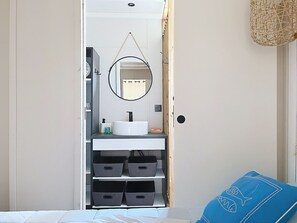 Une chambre à coucher avec un lit, un oreiller bleu, une table de chevet blanche, une commode blanche avec un miroir et un lavabo blanc.