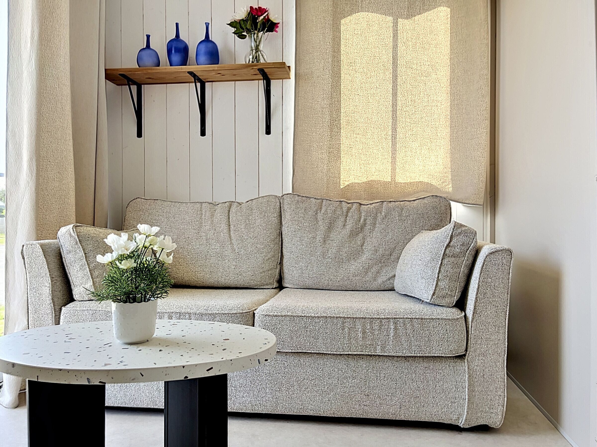 Een grijze bank met een witte kussen, een ronde tafel met een potplant, en een plank met blauwe vazen en een vaas met bloemen.