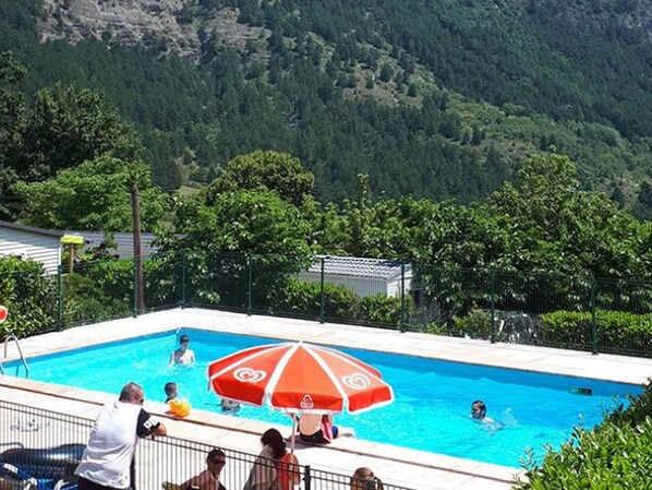 Ein Swimmingpool mit rotem Sonnenschirm, umgeben von viel Grün und Bergen im Hintergrund.