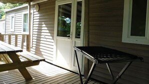 Katettu ulkotila, jossa on puinen terassi, metallipöytä ja ovi, joka johtaa ikkunoilla varustettuun rakennukseen.