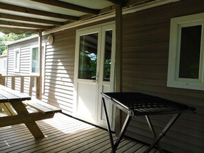 Ein überdachter Außenbereich mit Holzterrasse, einem Metalltisch und einer Tür zu einem Gebäude mit Fenstern.