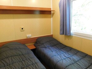Ein kleines Zimmer mit zwei Einzelbetten, einem Holzregal, einem Fenster mit Vorhängen und einer Steckdose.