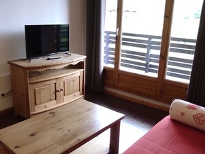 Ein Holz-TV-Möbel mit Flachbildfernseher, ein kleiner Tisch, ein rotes Sofa und ein Fenster mit Blick auf eine Holzterrasse.