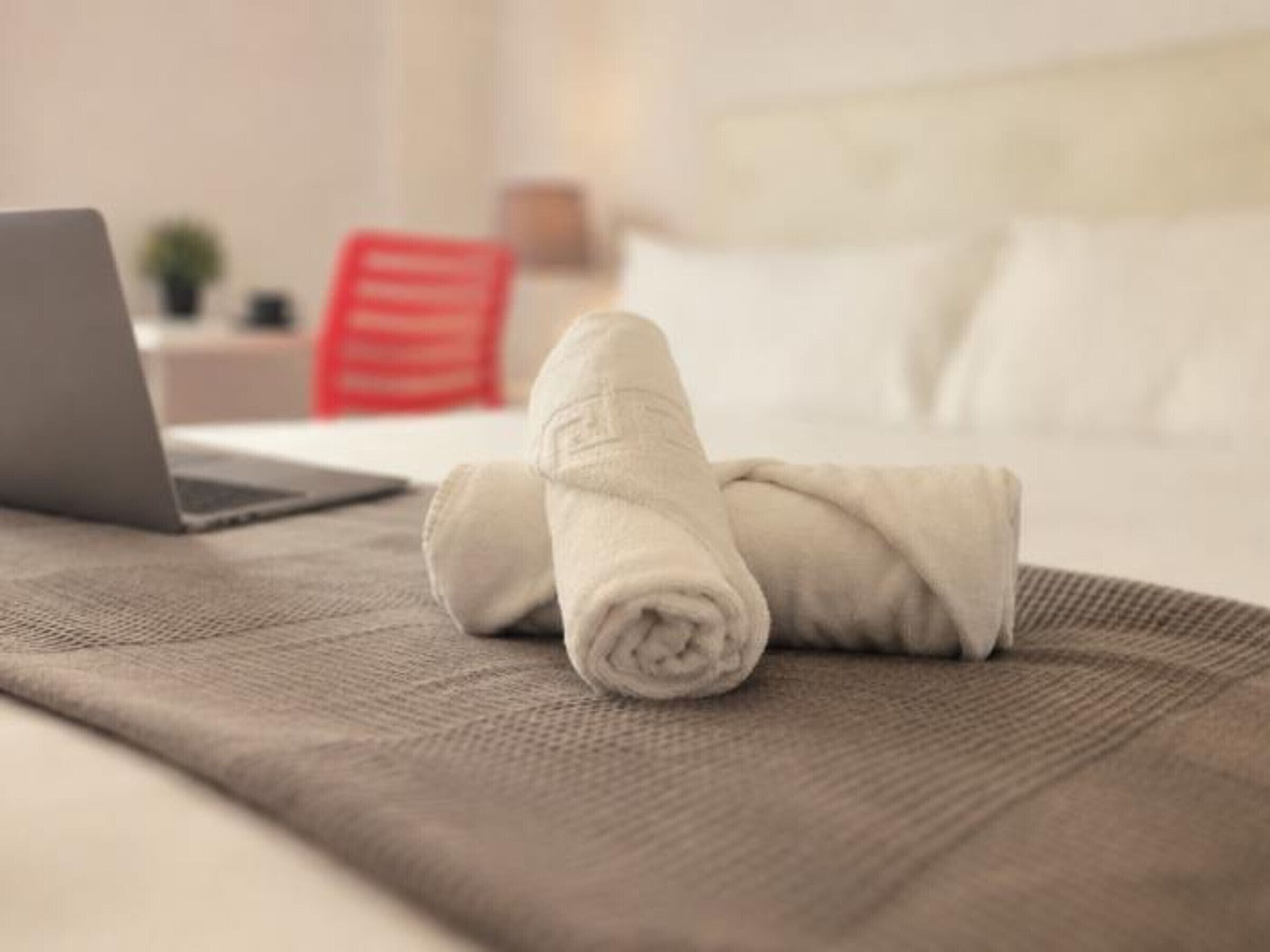 A neatly made bed with a laptop on a textured blanket, a red chair, and a white pillow in the background.