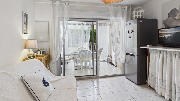 A room with a bed, a TV, a refrigerator, a dining area with chairs, and a view through a glass door. - Studio with pool, parking - 4 guests (Marseillan-Plage)