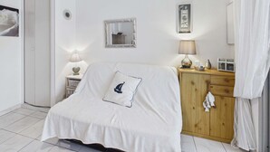 A bedroom with a bed covered in a white blanket, a wooden dresser, a mirror, and a bedside table with a lamp. - Studio with pool, parking - 4 guests (Marseillan-Plage)