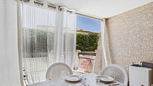 A dining area with a table set for two, a chair, a small table, and a white cabinet. - Studio with pool, parking - 4 guests (Marseillan-Plage)
