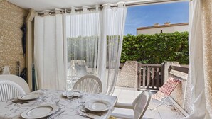 A dining area with a table set for four, chairs, and a view of a garden and building through a glass door. - Studio with pool, parking - 4 guests (Marseillan-Plage)