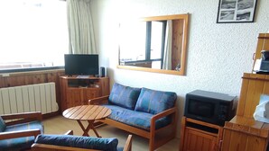 Ein wohnlich eingerichtetes Wohnzimmer mit Sofa, Sesseln, einem kleinen Tisch, einem Fernseher und einem Schrank.