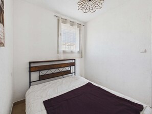 Ein Einzelbett mit Holz Kopfteil, ein Fenster mit leichtem Vorhang und eine schlichte weiße Wand.