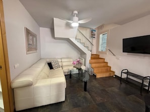 Ein Wohnzimmer mit einem weißen Ledersofa, einem Glastisch, einem weißen Deckenventilator, einer Treppe und einem an der Wand montierten Flachbildfernseher.