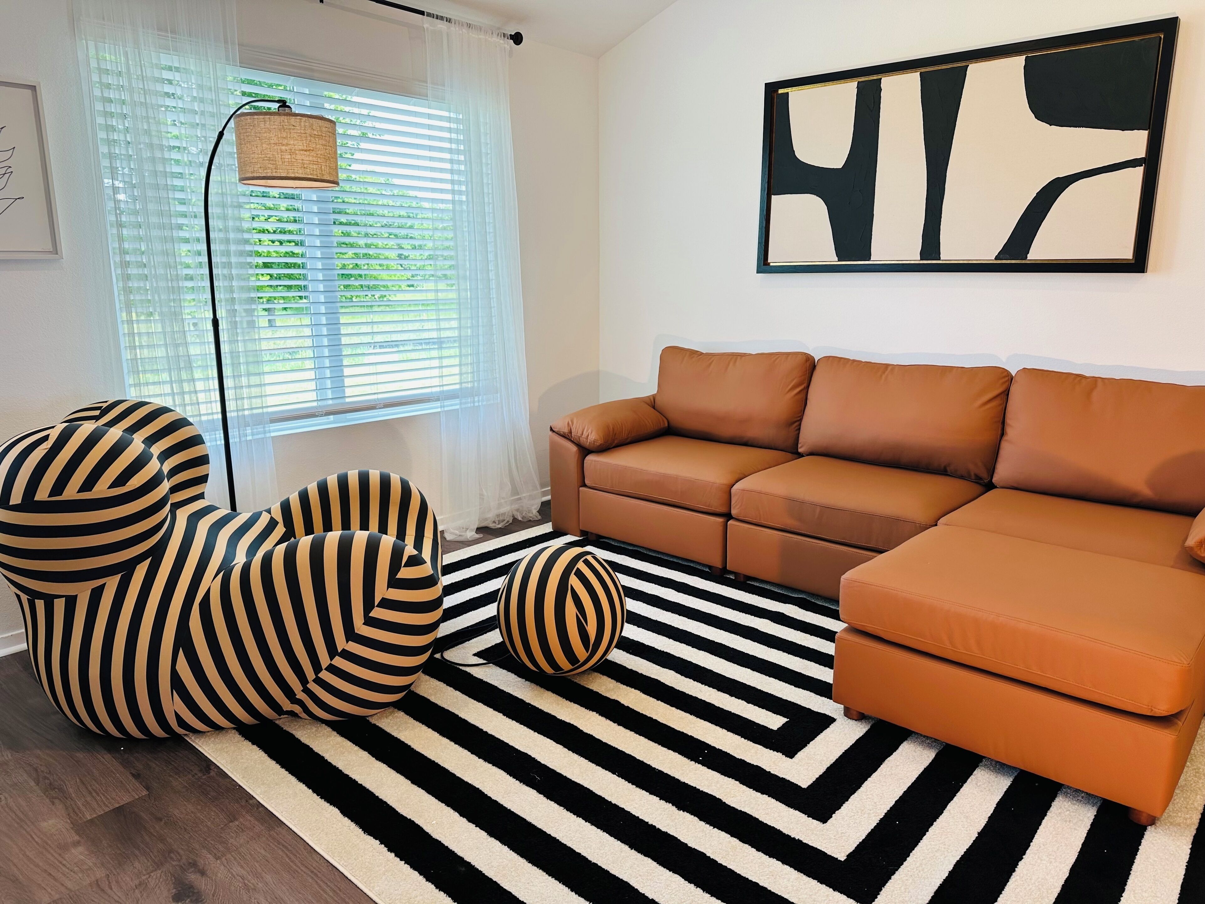 A living room with a striped sofa, a striped armchair, a round ottoman, a black and white striped rug, a floor lamp, and a large abstract painting on the wall.. Green Pastures - Another Blessing House
