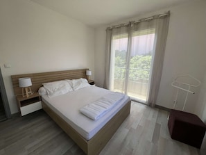 A bedroom with a wooden bed, a nightstand with a lamp, a window with curtains, and a small stool. - Apartment in city center, small sea view - 4 guests (Valras-Plage)