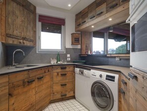 A kitchen with wooden cabinets, a built-in washing machine, and a window with a red blind. - Ski-in/Ski-out apartment - 4 guests (Courchevel 1850)