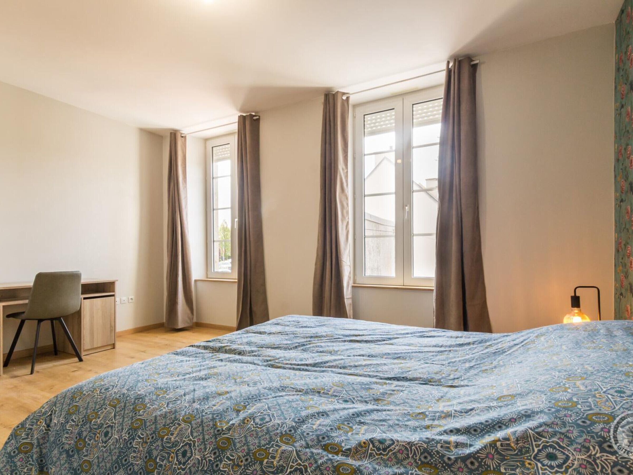 Ein Schlafzimmer mit einem Bett, einem Schreibtisch und zwei mit Vorhängen versehenen Fenstern.