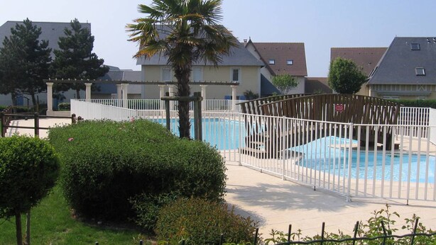 A swimming pool with a white fence, surrounded by greenery and residential buildings. - Cottage with garden and pool - 4 guests (Dives-sur-Mer)