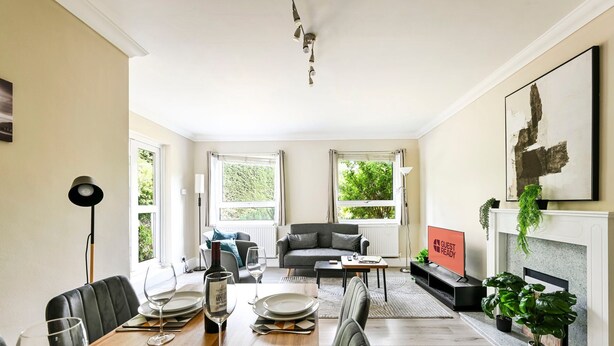 A dining area with a wooden table set for four, a grey sofa, and a fireplace with a 'GUEST READY' sign. - GuestReady - Comfortable 2bed in Richmond (London)