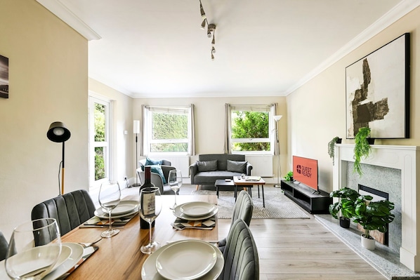 Ein Essbereich mit einem Holztisch, gedeckt für vier Personen, einer grauen Couch und einem Kamin mit dem Schild 'GUEST READY'.