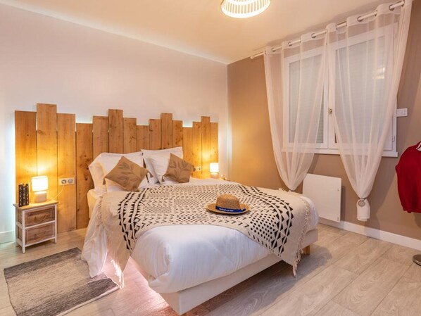 A bedroom with a wooden headboard, a bed with patterned bedding, a nightstand with a lamp, and a window with sheer curtains.