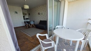 Un balcón con una mesa y sillas blancas, una puerta corrediza de vidrio, una zona de cocina y una sala de estar con sofá y sillones.
