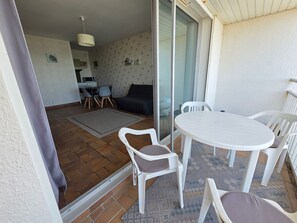 A balcony with a white table and chairs, a sliding glass door, a kitchen area, and a living room with a sofa and chairs. - Apartment with port view - 4 guests (Jard-sur-Mer)