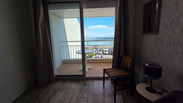 Una habitación con puerta corrediza de vidrio que da a un balcón, una mesita con una silla, un televisor sobre un mueble, y vista al mar y a los barcos.