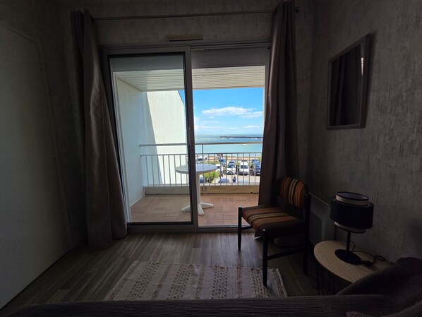 A room with a sliding glass door leading to a balcony, a small table with a chair, a TV on a stand, and a view of the sea and boats. - Apartment with port view - 4 guests (Jard-sur-Mer)