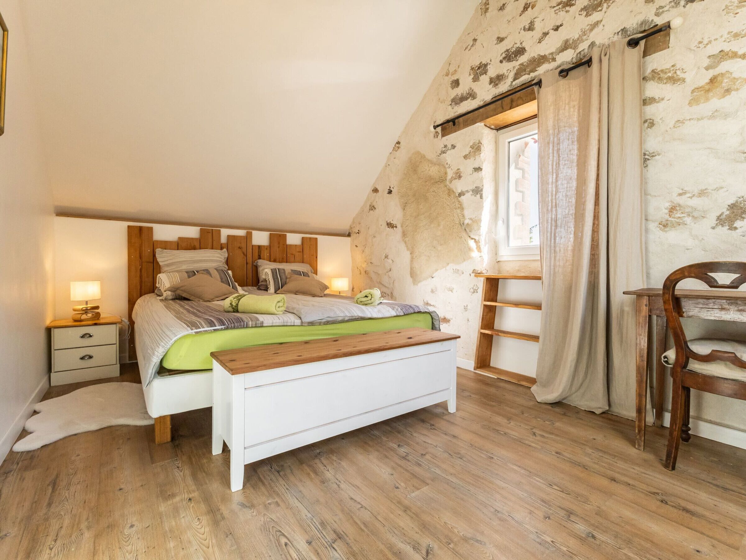A bedroom with a wooden headboard, a bed with striped bedding, a bedside table with a lamp, a wooden bench, and a chair.
