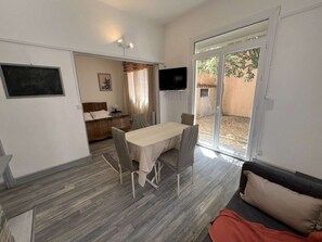 A room with a dining table, chairs, a sofa, a TV, and a sliding glass door leading to an outdoor area. - Charming T2 city center, garden - 2 guests (Valras-Plage)