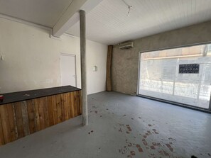 A room with a wooden counter, a white door, a concrete pillar, and a large window with a view of a street. - Charming T2 city center, garden - 2 guests (Valras-Plage)