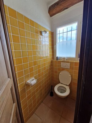 Een kleine badkamer met gele tegels, een toilet, een raam en een douche.