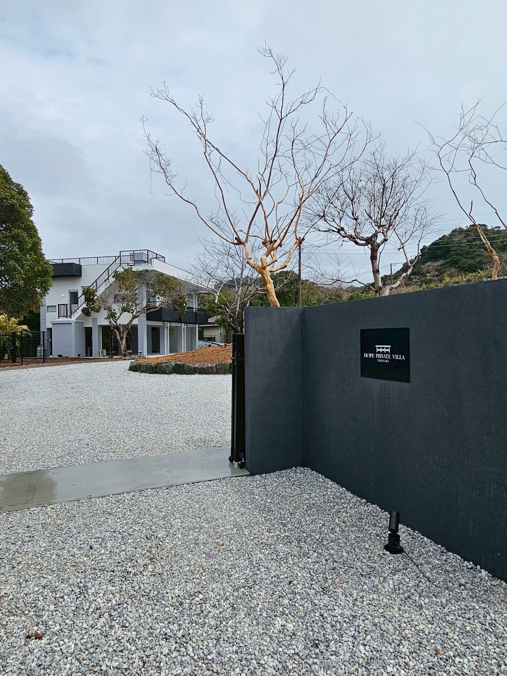 A gravel driveway leading to a modern building with a black fence and a sign indicating 'HOPE PRIVATE VILLA.'