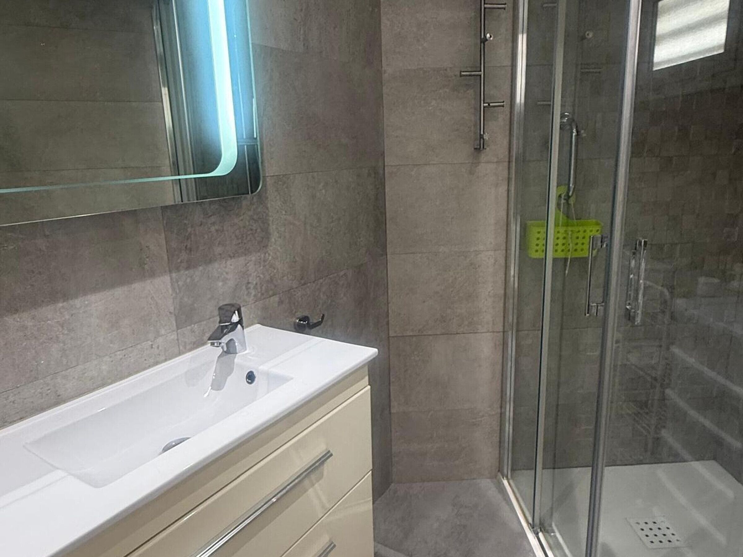 A bathroom with a white sink, a mirror, a glass shower enclosure, and a green cleaning basket.