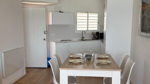 Een eetruimte met een tafel voor vier personen, witte stoelen, een keuken op de achtergrond en een deur naar een andere kamer.