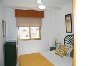 Ein Schlafzimmer mit einem Bett, einem Deckenventilator, einem Fenster mit Jalousien, einem Nachttisch und einer Wandlampe.