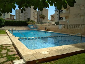Ein rechteckiger Pool mit einer gefliesten Kante, umgeben von einem gepflasterten Gehweg, neben einem Gebäude mit mehreren Balkonen.